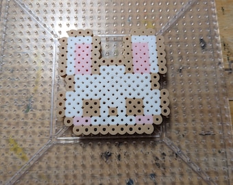 Può includere: Uno sprite di coniglio di perline bianche con orecchie rosa e accenti marroni su una bacheca di plastica trasparente.