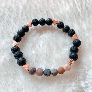 May include: A black lava rock bracelet with rose gold heart charms and a row of six natural stone beads in shades of brown, grey, and pink.