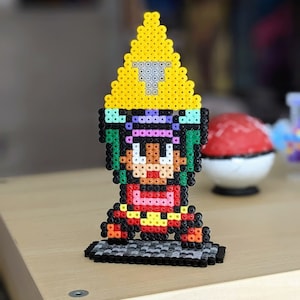 Könnte beinhalten: Eine Pixel-Art-Figur eines Charakters, der einen gelben Hut, ein lila-rosa Hemd und eine rote Hose trägt. Die Figur ist aus bunten Plastikperlen gefertigt und steht auf einem schwarzen Sockel.