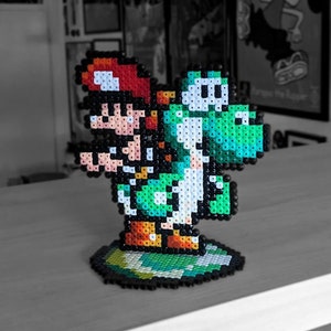 May include: A green Yoshi and Mario character made of perler beads. The characters are standing on a green and brown base.