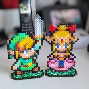 Könnte beinhalten: Eine grüne und gelbe Pixel-Art-Figur von Link aus der Videospielserie The Legend of Zelda, die ein Schwert hält, steht neben einer pinkfarbenen und gelben Pixel-Art-Figur von Prinzessin Zelda. Beide Figuren stehen auf grünen Sockeln.
