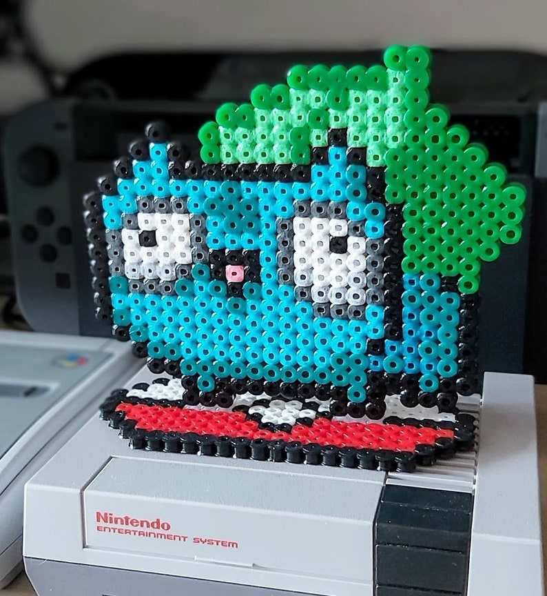 Puede incluir: Una figura de pixel art verde, azul y blanca de un personaje de un videojuego. La figura est&aacute; de pie sobre una plataforma roja y negra. La figura est&aacute; encima de una consola Nintendo Entertainment System.