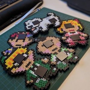 Pu&ograve; includere: Cinque magneti colorati in pixel art che rappresentano personaggi con diversi stili di capelli e vestiti. Ogni magnete ha un quadrato nero sul retro per essere attaccato a una superficie metallica.
