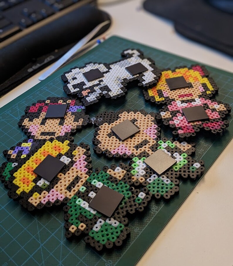 Pu&ograve; includere: Cinque magneti colorati in pixel art che rappresentano personaggi con diversi stili di capelli e vestiti. Ogni magnete ha un quadrato nero sul retro per essere attaccato a una superficie metallica.
