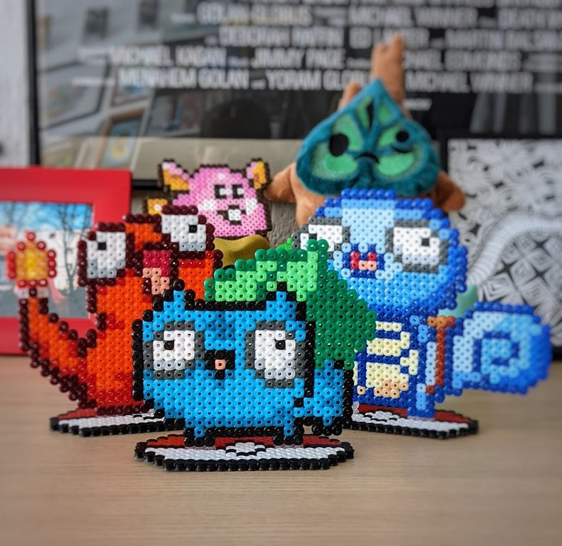Puede incluir: Cuatro figuras de arte pixelado de colores hechas de cuentas. Las figuras son una criatura azul y verde, una criatura azul con un pico amarillo, una criatura roja con una llama amarilla y una criatura rosa con una estrella amarilla. Las figuras est&aacute;n todas de pie sobre una superficie de madera.