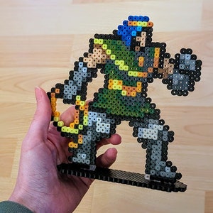 May include: A green and grey pixel art figure of a warrior holding a sword and shield. The figure is made of plastic beads and is standing on a black base.