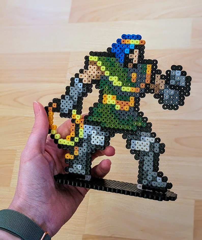 May include: A green and grey pixel art figure of a warrior holding a sword and shield. The figure is made of plastic beads and is standing on a black base.
