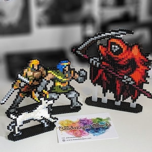 Könnte beinhalten: Drei Pixel-Art-Figuren aus bunten Perlen. Die Figuren sind eine rote, mit einer Sense ausgestattete Figur mit Kapuze, eine grüne und gelbe Kriegerfigur und eine gelbe Kriegerfigur mit einem weißen Hund. Die Figuren stehen auf schwarzen Sockeln. Die Figuren wurden von Beaddazepin Works hergestellt.