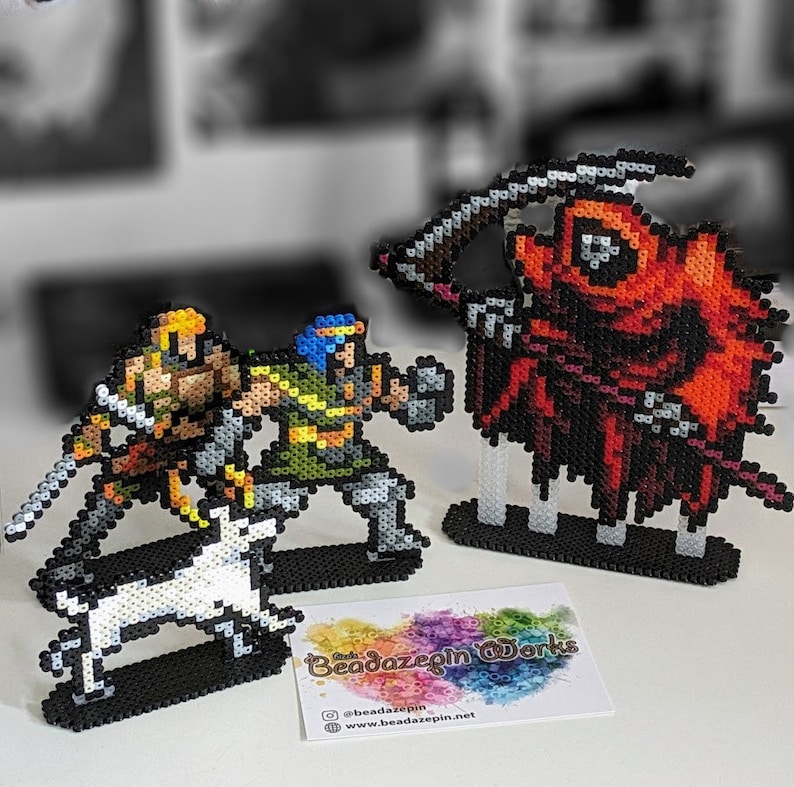 May include: Three pixel art figures made of colourful beads. The figures are a red hooded figure with a scythe, a green and yellow warrior figure, and a yellow warrior figure with a white dog. The figures are on black bases. The figures are made by Beaddazepin Works.