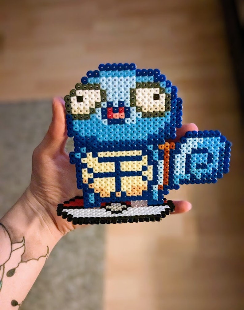 Puede incluir: Un personaje de pixel art azul y blanco con un caparaz&oacute;n en la espalda, ojos grandes y la lengua fuera. El personaje est&aacute; de pie sobre una plataforma roja y blanca.