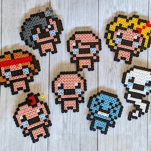 May include: A collection of pixel art figures, each depicting a crying character with different hair colors and accessories. The figures are made from small, colorful beads and arranged on a light wood-grain surface. The characters have black outlines and blue tear drops.