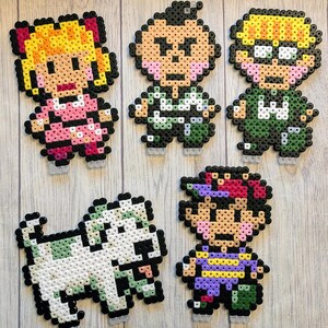 Pu&ograve; includere: Cinque personaggi in pixel art e un cane realizzati con perline Perler colorate. I personaggi includono una ragazza con i capelli biondi e un vestito rosa, un ragazzo con i capelli castani e una camicia verde, un ragazzo con i capelli biondi e una camicia verde, un ragazzo con i capelli castani e una camicia viola, e un cane bianco.