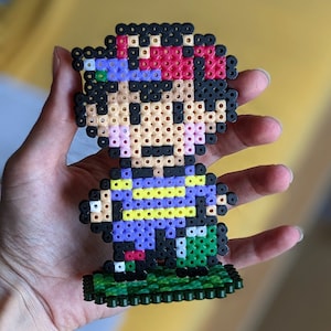 Pu&ograve; includere: Una figura di pixel art di un personaggio di videogioco realizzata con perline colorate. Il personaggio indossa una camicia viola, pantaloni neri e un cappello rosso. La figura &egrave; in piedi su una base verde.
