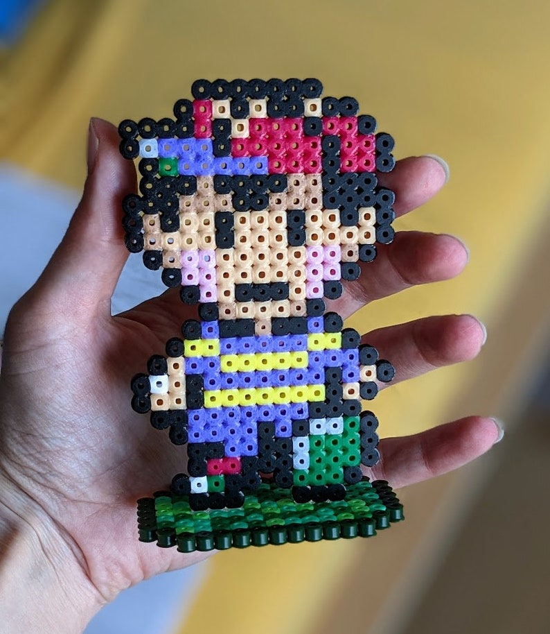 Pu&ograve; includere: Una figura di pixel art di un personaggio di videogioco realizzata con perline colorate. Il personaggio indossa una camicia viola, pantaloni neri e un cappello rosso. La figura &egrave; in piedi su una base verde.