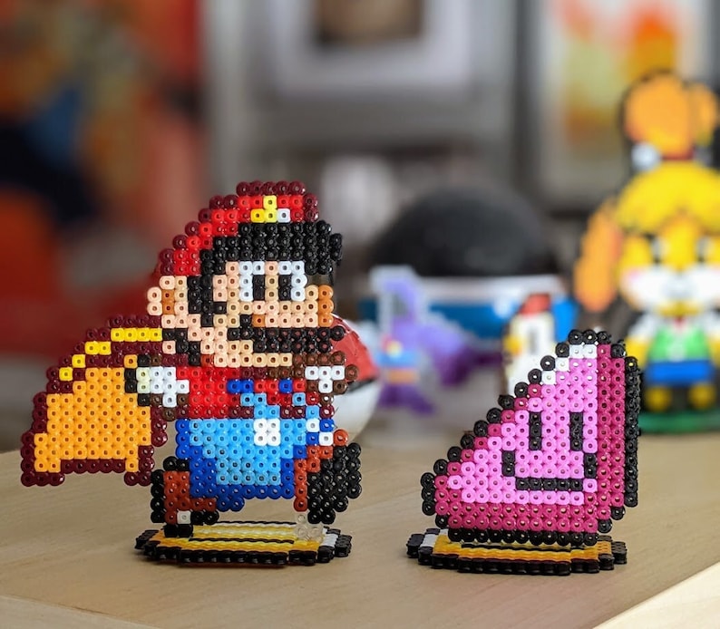 May include: A red and black Super Mario character made of perler beads. The character is holding a yellow and brown object. A pink and white perler bead character is in the background.