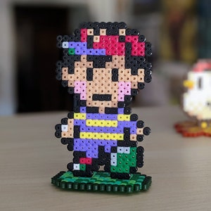 Pu&ograve; includere: Una figura di pixel art di un personaggio con capelli neri, un cappello rosso e viola, una camicia viola e gialla e pantaloni verdi in piedi su una base verde.