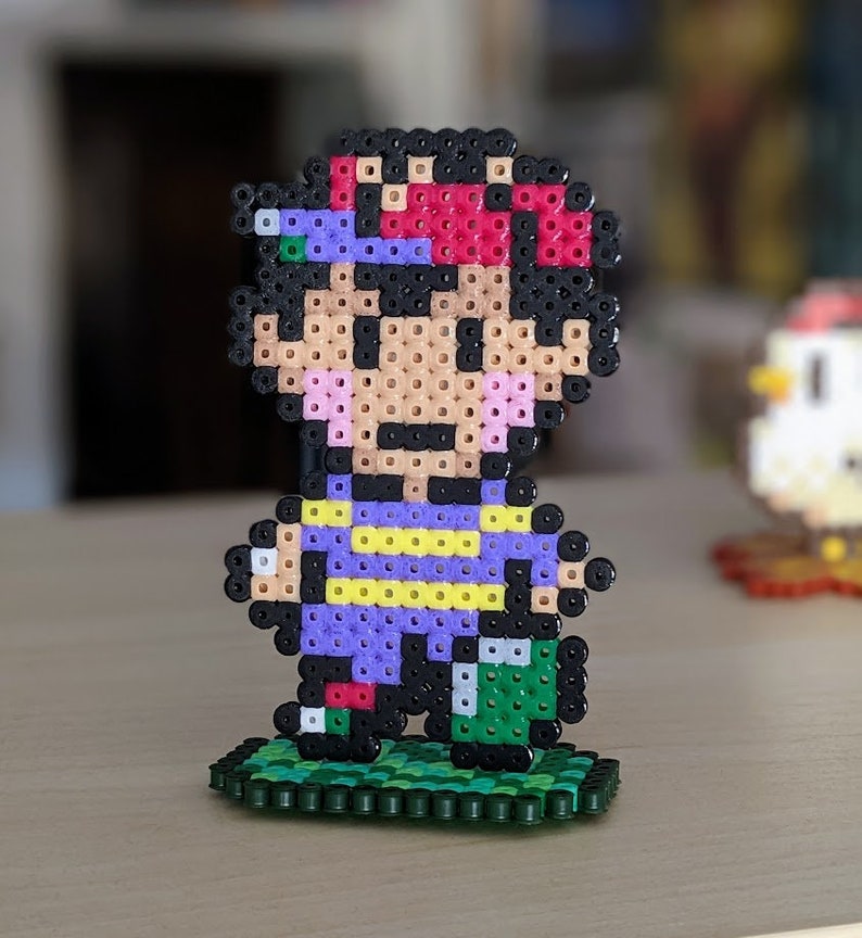 Pu&ograve; includere: Una figura di pixel art di un personaggio con capelli neri, un cappello rosso e viola, una camicia viola e gialla e pantaloni verdi in piedi su una base verde.