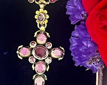 May include: A gold-toned pendant necklace with a cross design, featuring oval and round pink gemstones. The necklace is suspended from a delicate chain. The background includes purple flowers and a red rose.