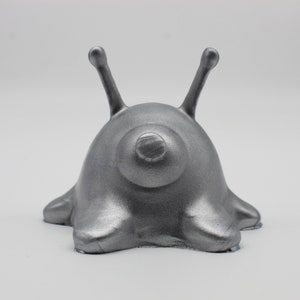 May include: A silver-colored alien figurine with two antennas and a round body. The figurine is sitting on a white surface.