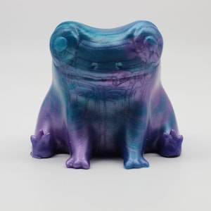 May include: A small, iridescent purple and blue frog figurine with a glossy finish. The frog is sitting with its legs crossed and has a wide, toothy grin.