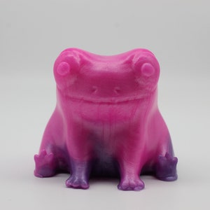 May include: A small, pink and purple frog figurine with a glossy finish. The frog is sitting with its legs crossed and has a wide smile.