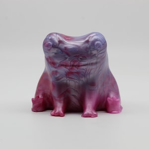 May include: A small, hand-painted ceramic frog figurine with a pink and purple iridescent glaze. The frog is sitting with its legs tucked underneath it.