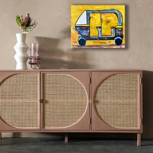 May include: An abstract painting of a yellow and blue auto-rickshaw with the name "Sonya" on the side. The artwork hangs above a mid-century modern cabinet with woven cane door panels and a vase with a flower.