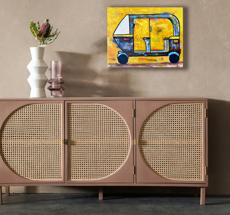 May include: An abstract painting of a yellow and blue auto-rickshaw with the name "Sonya" on the side. The artwork hangs above a mid-century modern cabinet with woven cane door panels and a vase with a flower.