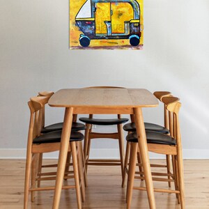 May include: An abstract painting of a yellow and black auto-rickshaw on a yellow background. The artwork is displayed above a wooden dining table with six chairs. The chairs have black seats and light wood frames.