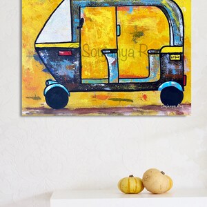 May include: An abstract painting of an auto-rickshaw, or tuk-tuk, in shades of yellow, blue, and black. The artwork features the text "Soujanya Rao" in the lower right corner. Two small pumpkins sit on a white shelf below the painting.