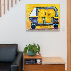 May include: An abstract painting of a yellow, blue, and white auto-rickshaw. The artwork is displayed on a white wall. The painting has a textured appearance and is signed by the artist. The image also shows a wooden bench with a cushion, a plant, and books.