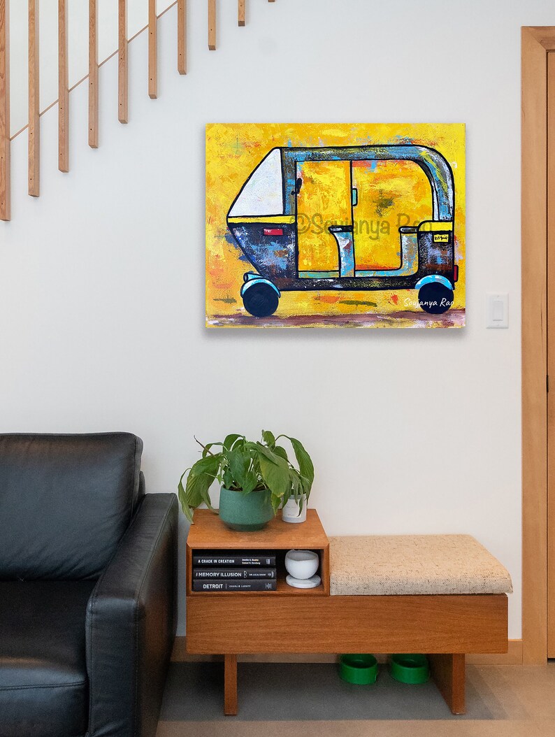 May include: An abstract painting of a yellow, blue, and white auto-rickshaw. The artwork is displayed on a white wall. The painting has a textured appearance and is signed by the artist. The image also shows a wooden bench with a cushion, a plant, and books.