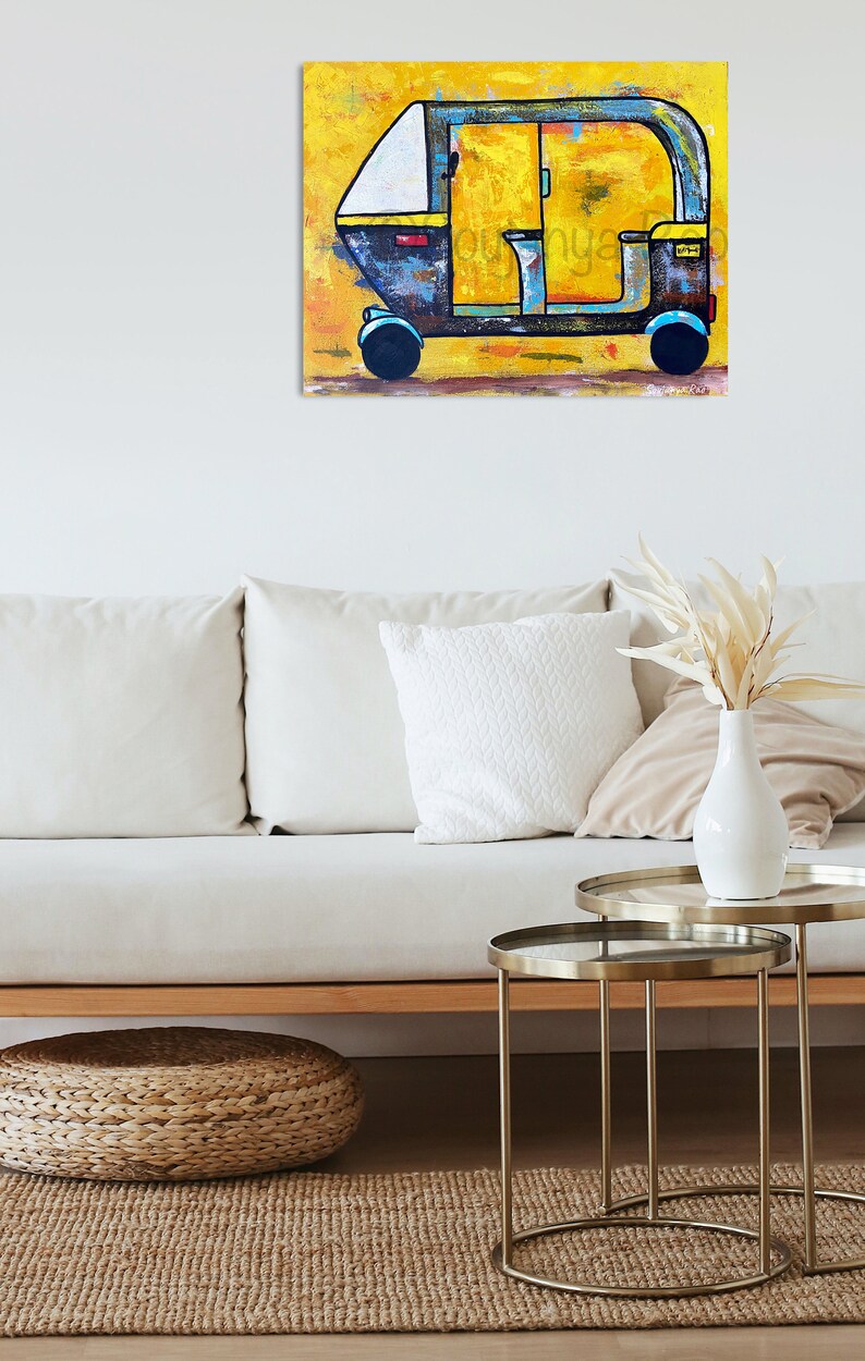 May include: A colorful painting of an auto rickshaw, or tuk-tuk, against a yellow background. The vehicle is primarily blue and yellow, with black wheels. The artwork is displayed on a wall above a white sofa and decorative tables.