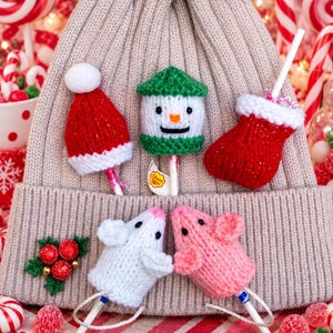 May include: A beige knit beanie with a faux fur pom-pom, decorated with five small knitted covers for lollipops. The covers include a red Santa hat, a snowman, a red stocking, and two mice in white and pink. A green holly sprig is attached.
