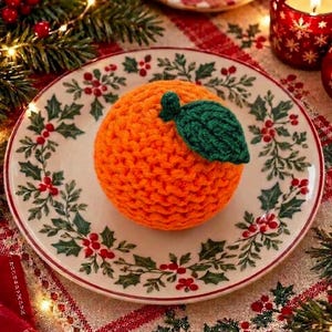 May include: A round, bright orange crocheted fruit ornament featuring a single dark green crocheted leaf attached to the top. The textured, yarn-based sphere is centered on a white ceramic plate decorated with a green holly and red berry pattern.