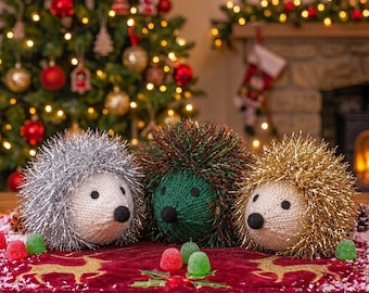 May include: Three plush hedgehog ornaments with knitted faces and tinsel quills in silver, green, and gold. Each features black felt eyes and a black nose. They are arranged on a red velvet runner decorated with gold reindeer and green gumdrops.