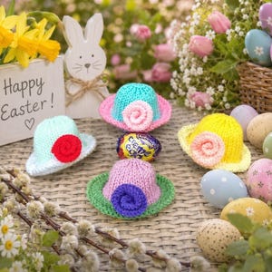 May include: Three small knitted hats in light blue, yellow, and lavender, each featuring a contrasting crocheted flower. The hats are arranged around a small chocolate egg, with a wooden bunny sign reading Happy Easter and various decorative eggs.