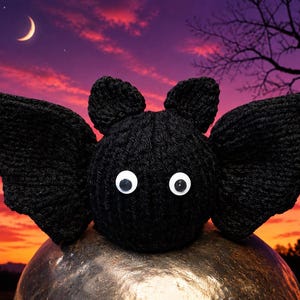 May include: A black crocheted bat plush toy featuring two pointed ears, large white googly eyes with black pupils, and wide, textured wings extending from the sides of its round, ribbed body, crafted with a consistent dark yarn texture.