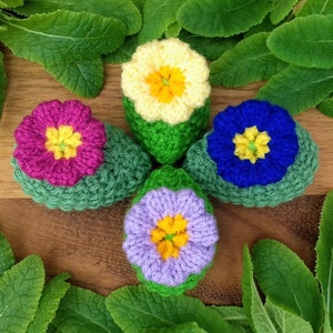 May include: Five crocheted flower-shaped ornaments in different colors, including pink, yellow, blue, and purple, with green crocheted stems. The ornaments are arranged in a circle on a wooden surface.