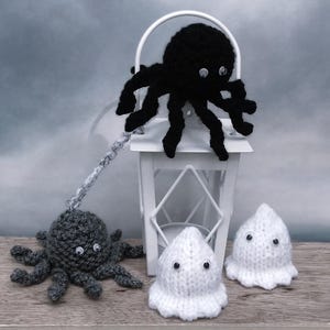 Puede incluir: Una exhibición temática de Halloween con criaturas de ganchillo hechas a mano. Un pulpo negro se encuentra sobre una linterna blanca, con un pulpo gris colgando de ella. También hay dos figuras de fantasmas blancos con ojos saltones, creando una escena espeluznante y festiva.