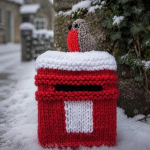 Könnte beinhalten: Ein handgestrickter Überzug für einen Briefkasten in Rot mit einem weißen oberen Teil, einem schwarzen horizontalen Schlitz und einem weißen rechteckigen Feld auf der Vorderseite. Ein kleiner gehäkelter braun-roter Rotkehlchen-Vogel sitzt obenauf.