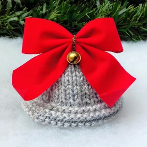 May include: A gray knitted hat with a red bow and a gold bell on top. The hat is sitting on a white fluffy surface.