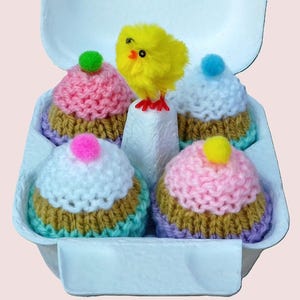 Könnte beinhalten: Vier gestrickte Oster-Eier in Cupcake-Form in einem weißen Eierkarton mit einem gelben Küken oben drauf. Die Cupcakes sind mit rosa, weißem und blauem Zuckerguss und Streuseln verziert.