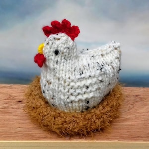 May include: A white knitted chicken with a red comb and yellow beak sitting in a brown faux fur nest.