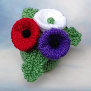Könnte beinhalten: Ein handgestricktes Blumenarrangement mit drei verschiedenen Blüten in Rot, Weiß und Violett, die jeweils eine kontrastierende Mitte aufweisen. Die Blüten sind an einer grünen Strickbasis mit strukturierten grünen Blättern befestigt und zeigen detaillierte Handarbeit.