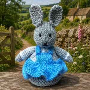 Puede incluir: Una muñeca de conejo tejida a mano de color gris con orejas largas y erguidas, ojos bordados en negro y una pequeña nariz negra. El conejo lleva un vestido tejido de color azul brillante con una falda con volantes y ribetes blancos.