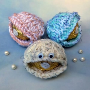 May include: Three handmade crochet candy holders, each shaped like an open shell. One is pink and peach, another is blue and teal, and the third is off-white with googly eyes. Each holds a gold-wrapped chocolate candy.