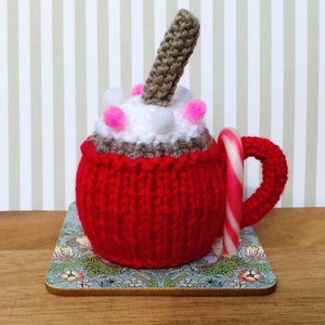 May include: A knitted red mug with a white and brown crocheted hot chocolate topping and a candy cane. The mug is sitting on a floral patterned coaster.