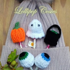 May include: A selection of Halloween-themed lollipop covers, including a knitted orange pumpkin, a white ghost, a black trick-or-treat basket, and two eyeball designs. The lollipops are displayed on a grey beanie with the text "Lollipop Cozies".