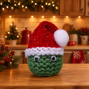 May include: A round, green, hand-knitted creature with two plastic googly eyes, wearing a red knitted hat with a white brim and a white pom-pom on top. The object has a textured, chunky yarn construction and sits on a flat wooden surface.